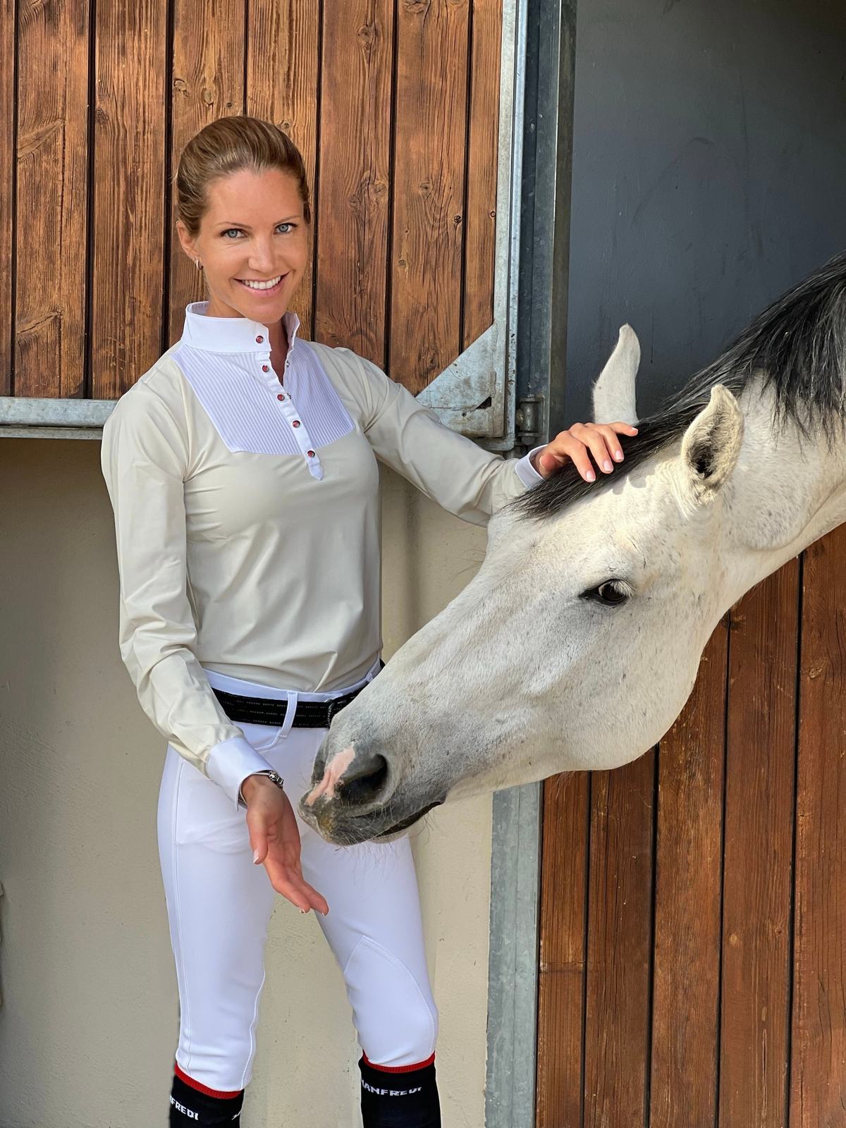 Diana sonriendo junto a la puerta del box mientras acaricia a un caballo tordo